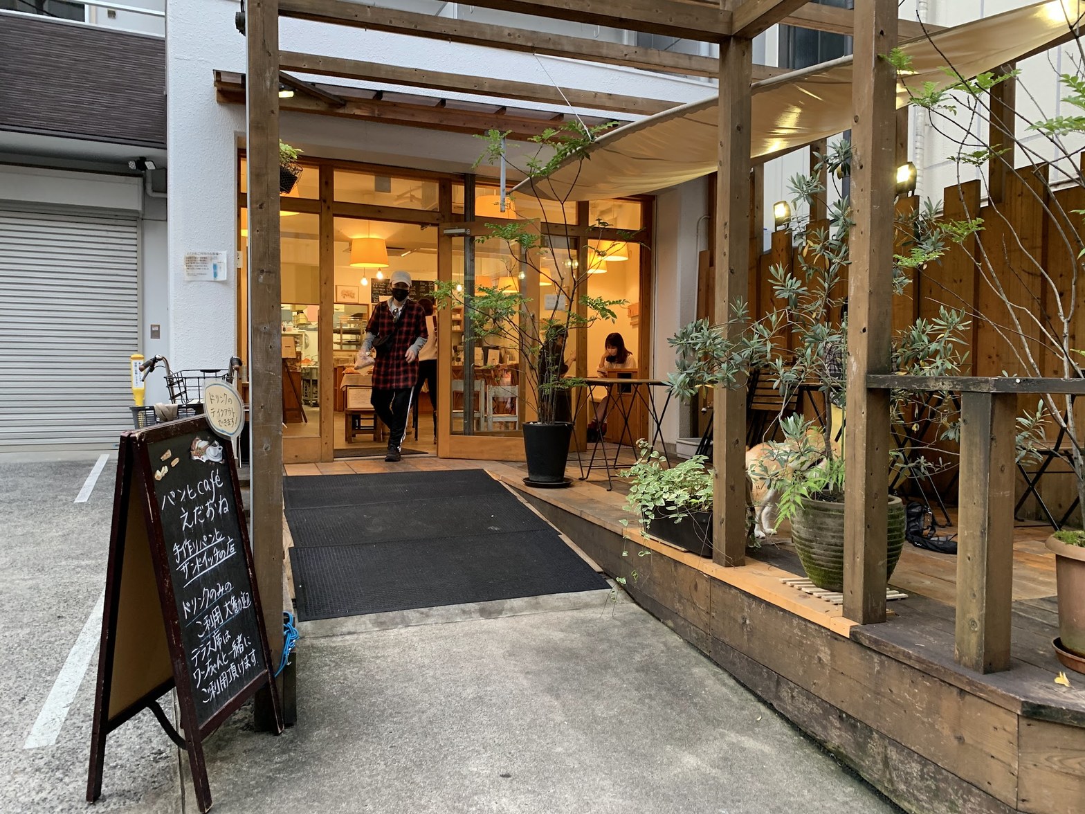 木製パーゴラとテラス席に囲まれた入口、黒板看板に「手作りパンとサンドイッチの店」の文字、店内に暖色の照明